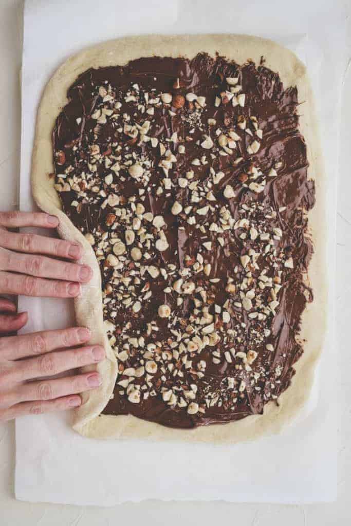 rolling up babka dough spread with nutella and topped with hazelnuts