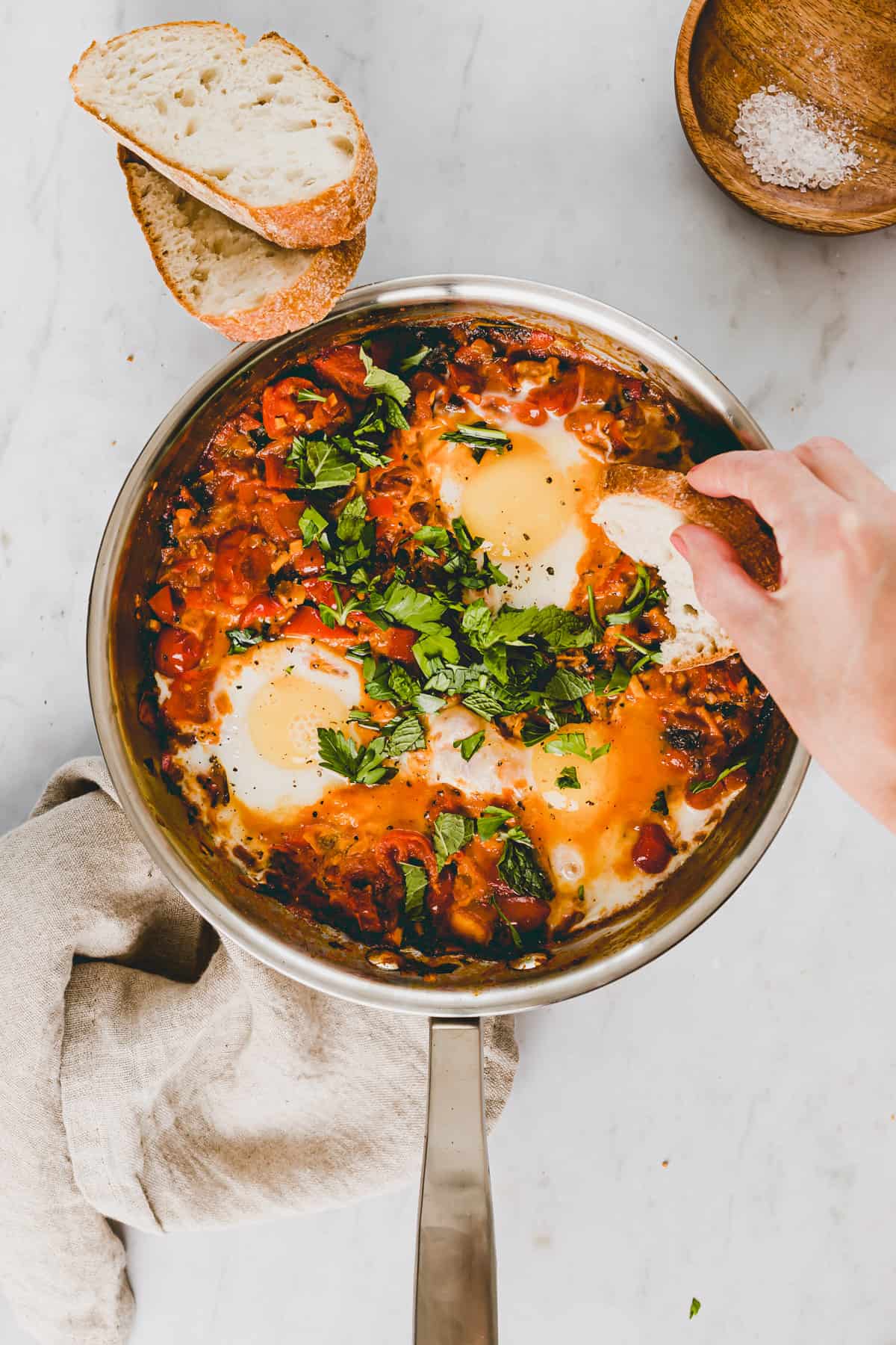 eine hand dippt ein stück brot in eine pfanne mit shakshuka