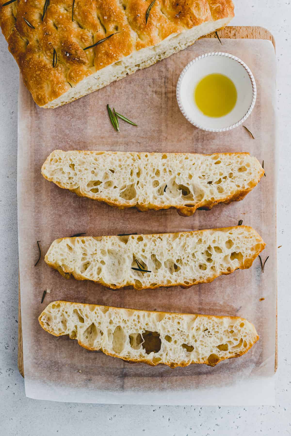 Top view of rosemary focaccia bread slices on a chopping board with a small bowl of olive oil on the side.  