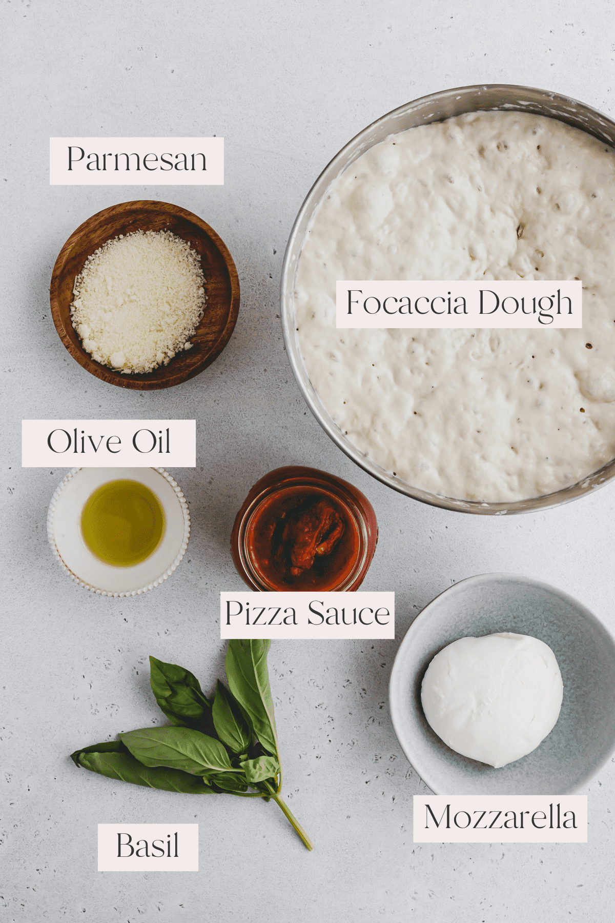 Top view of Focaccia Pizza Ingredients including focaccia dough, parmesan cheese, olive oil, pizza sauce, basil leaves and mozzarella. 