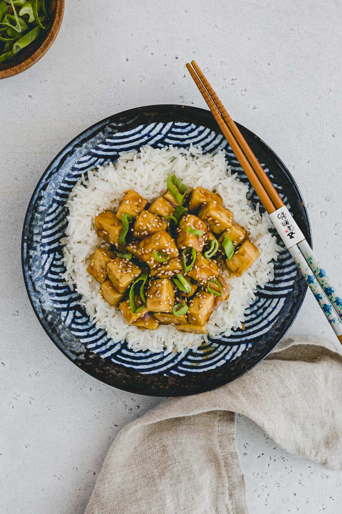 Top view and close up of teriyaki tofu on top of rice on a blue patterned plate with chopsticks on the side. 