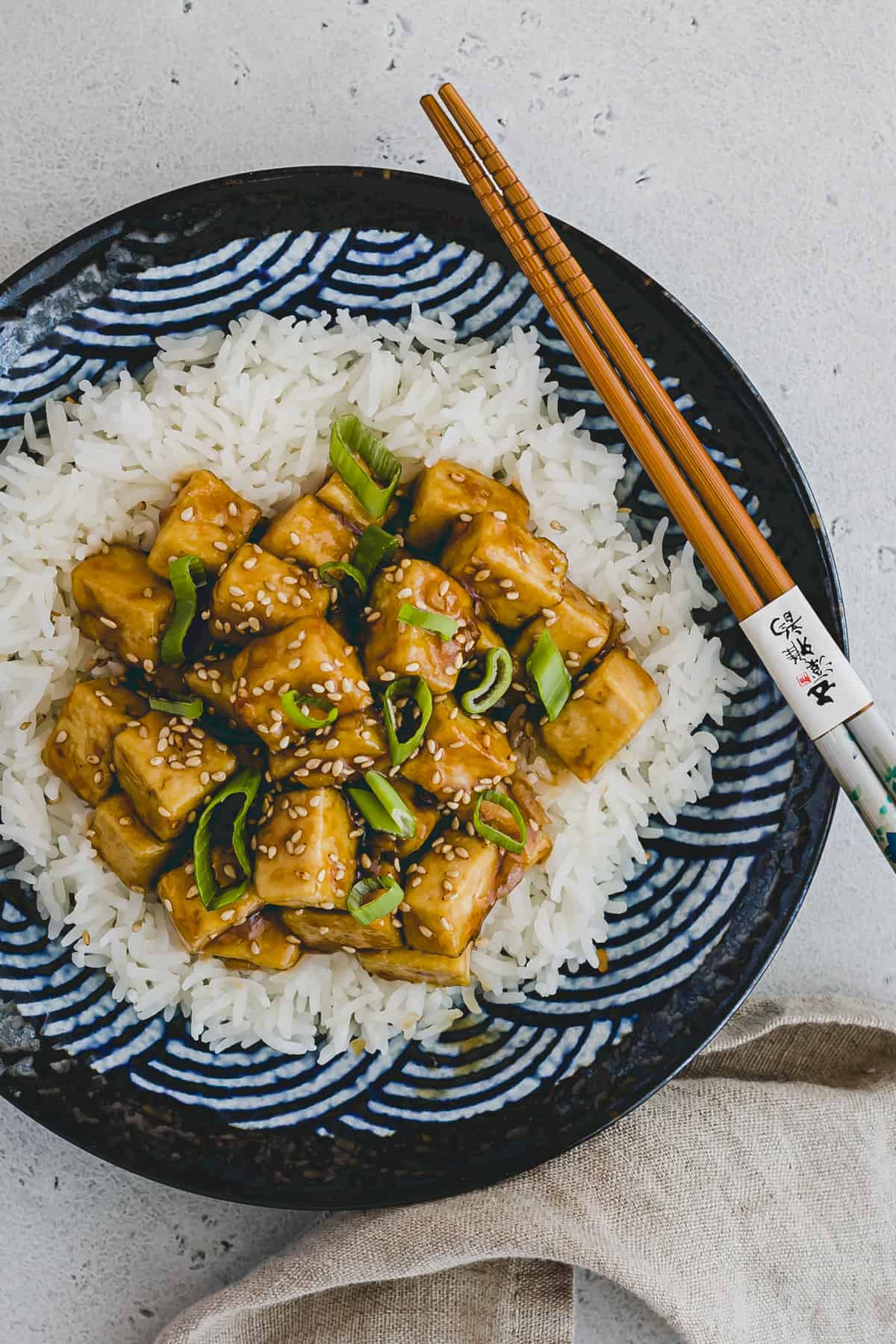 Top view and close up of teriyaki tofu on top of rice on a blue patterned plate with chopsticks on the side. 