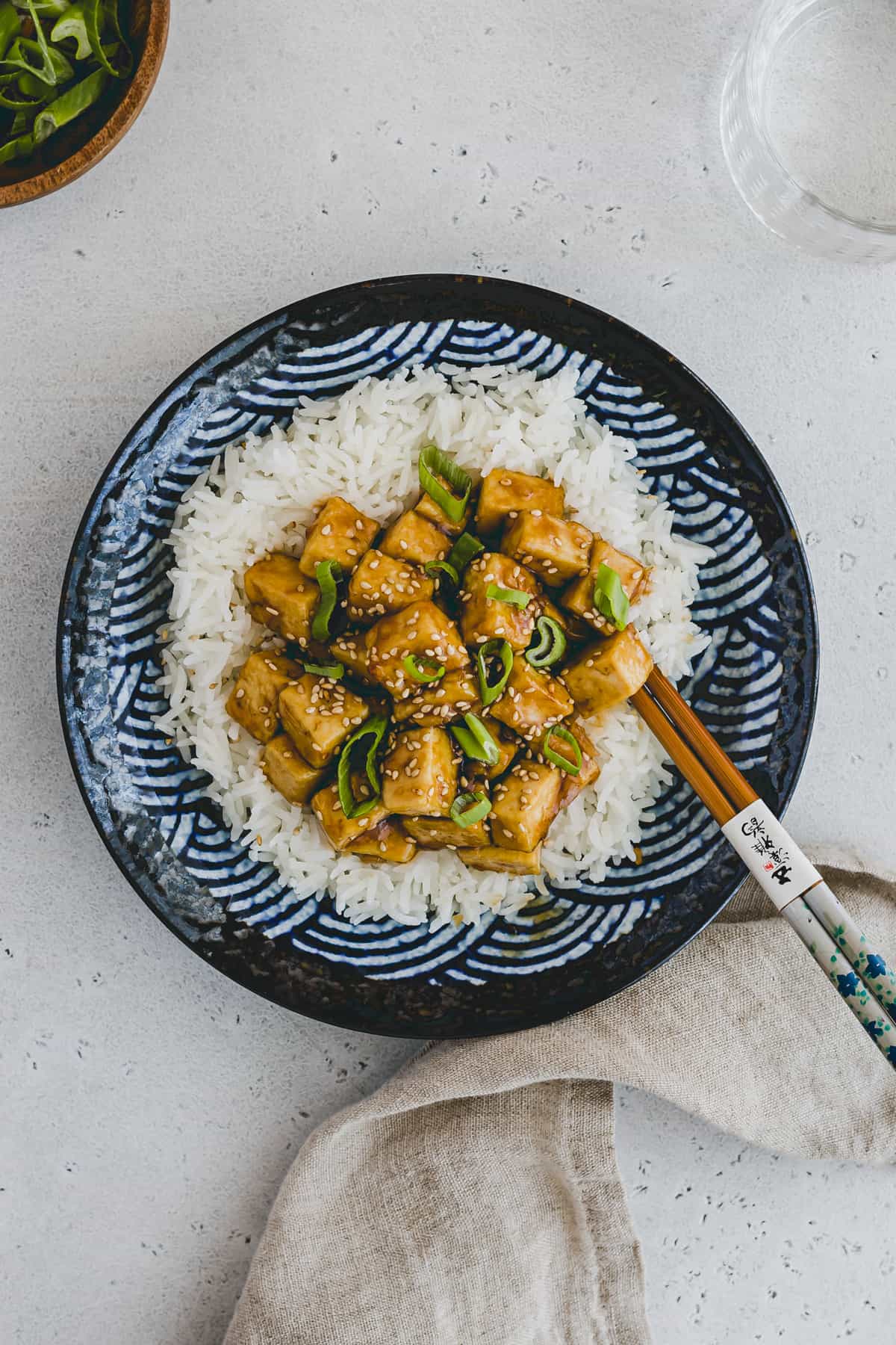 Top view of teriyaki tofu dish on top of rice on a blue patterned plate with chopsticks in the side. 