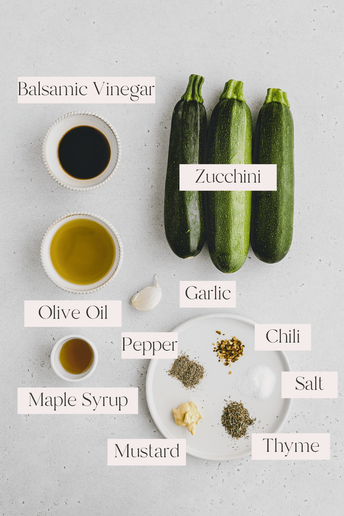 Top view of ingredients for grilled zucchini including zucchini, balsamic vinegar, olive oil, garlic clove, maple syrup, mustard, thyme, pepper, salt and red pepper flakes. 
