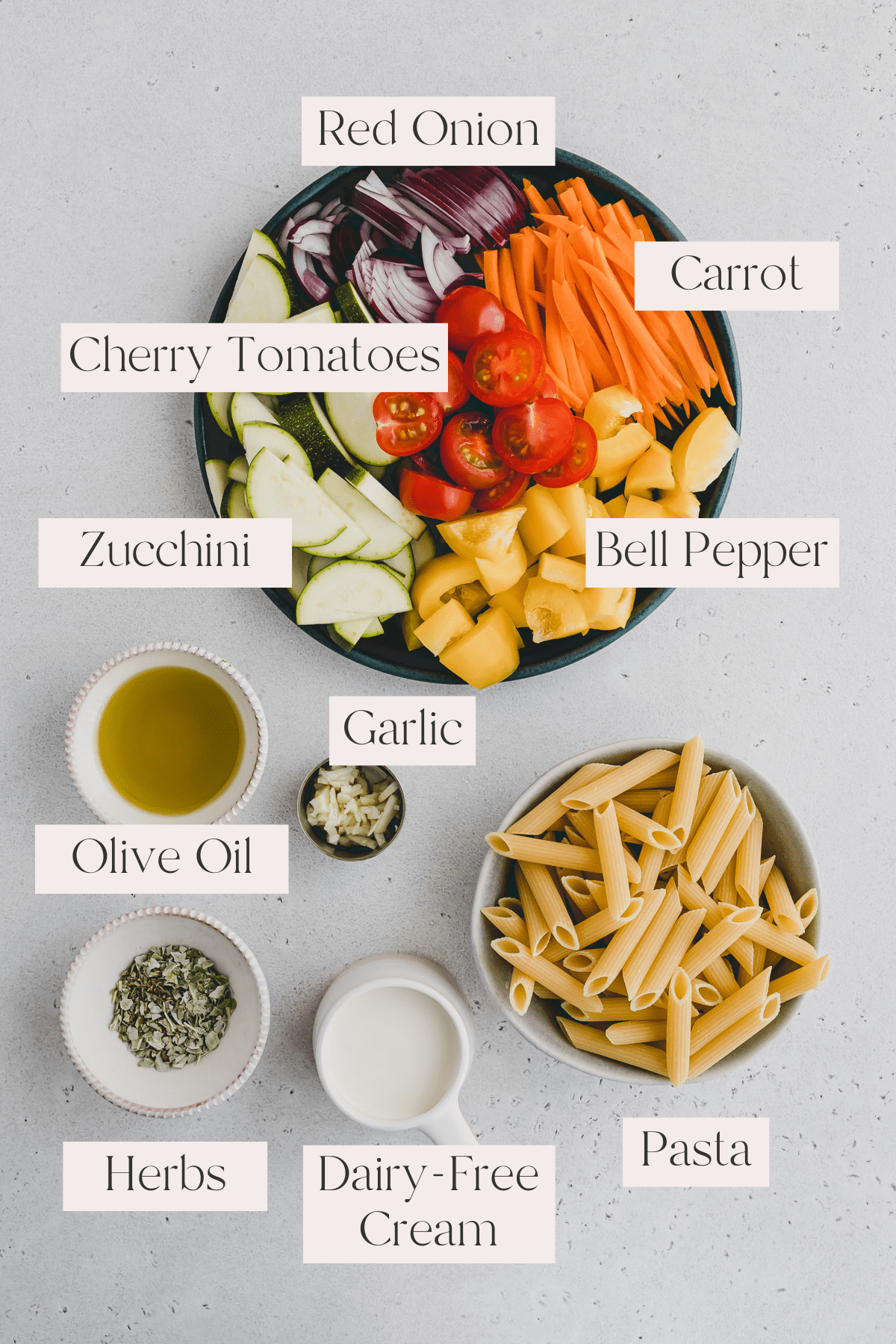 Top view of vegan Pasta Primavera Ingredients including a metal bowl filled with zucchini, bell pepper, red onion, cherry tomatoes and carrots,  and smaller bowls with garlic, olive oil, pasta, herbs, and dairy-free cream.