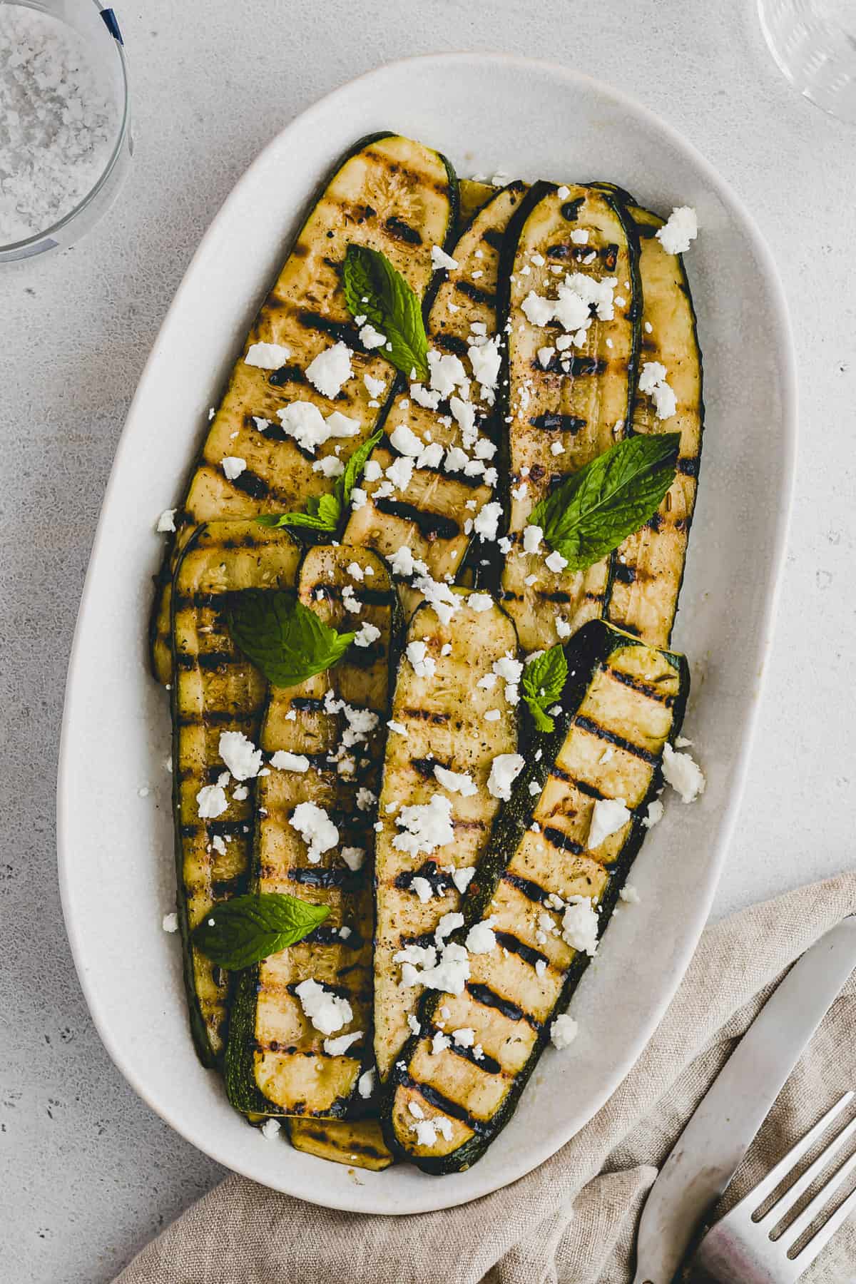 Top view of grilled slices of zucchini in a single layer on a white oval plate garnished with crumbled feta cheese and mint leaves. 