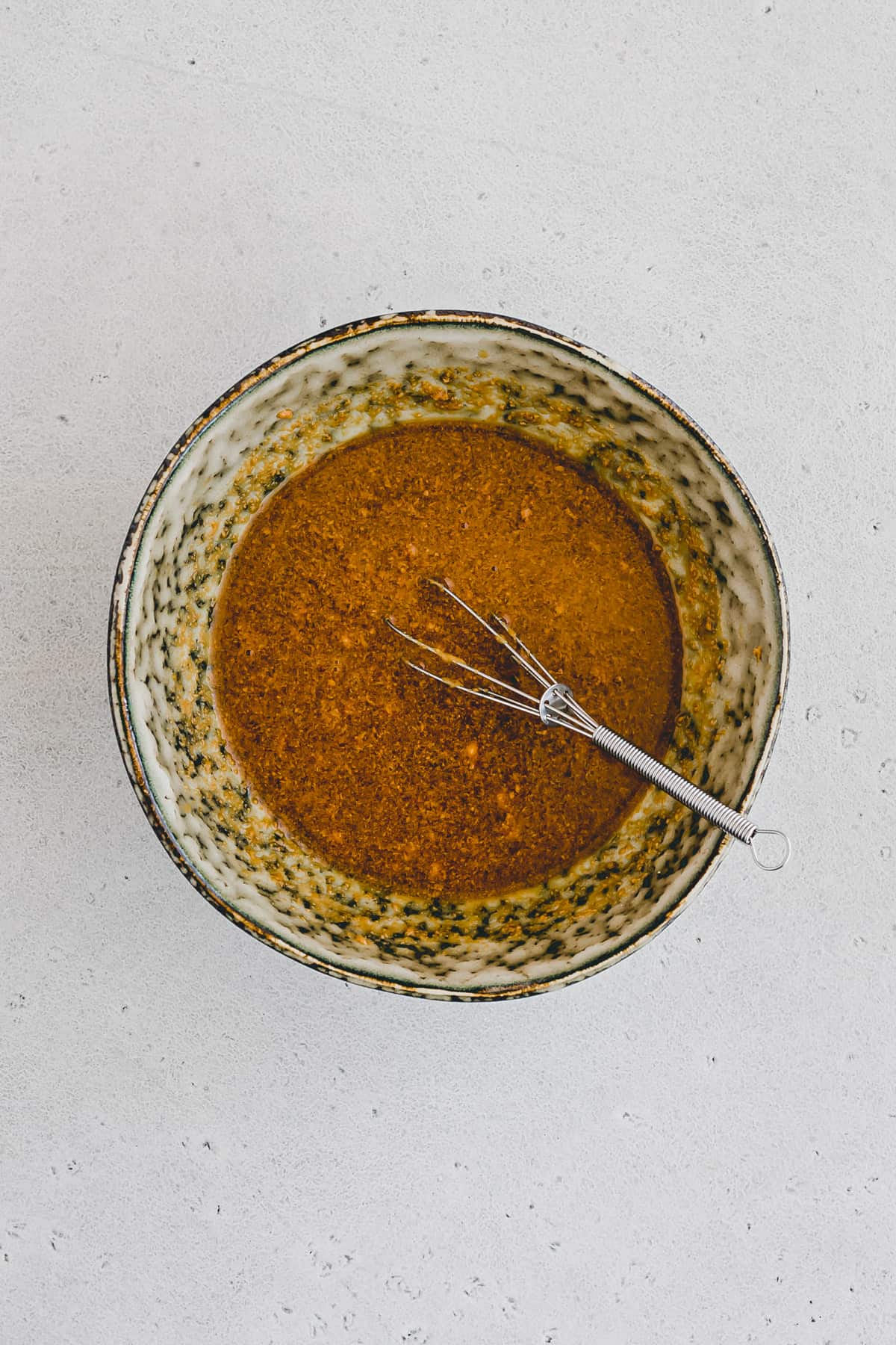 Top view close up shot of a brown spotted bowl with Asian salad dressing in it with a whisk in the bowl. 