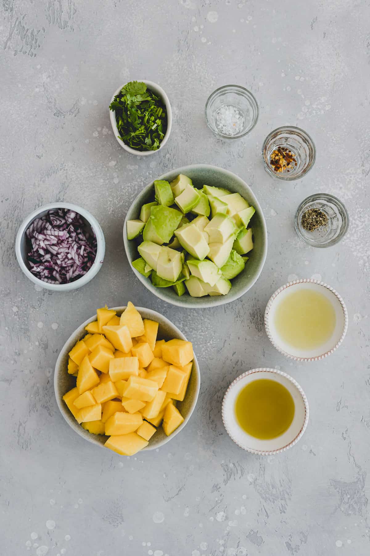 Top view of ingredients for mango avocado salad in small bowls, including diced mango, diced avocados, minced red onion, cilantro, olive oil, lime juice, salt & pepper, and red pepper flakes.