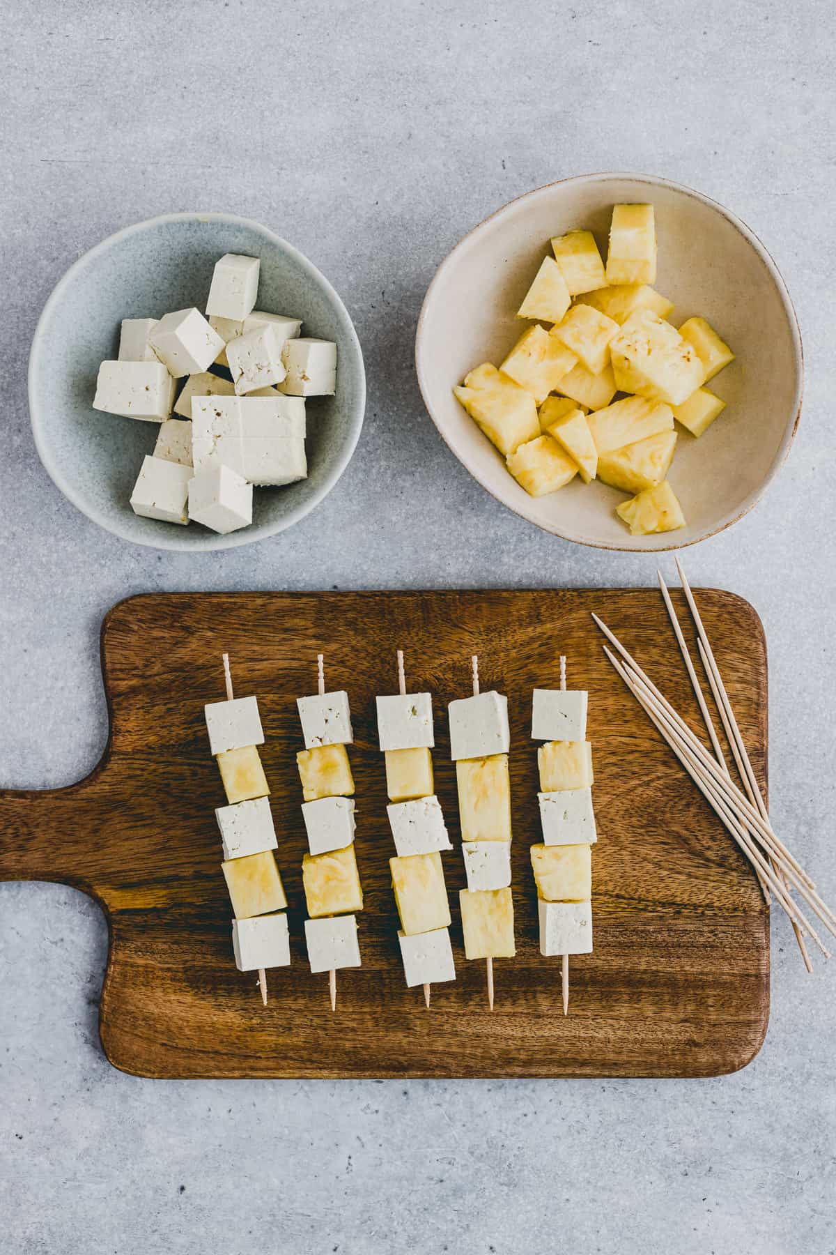 Top view of two bowls, one with cubes of tofu in it, the other with cubes of pineapple, with a chopping board of assembled tofu skewers next to it. 