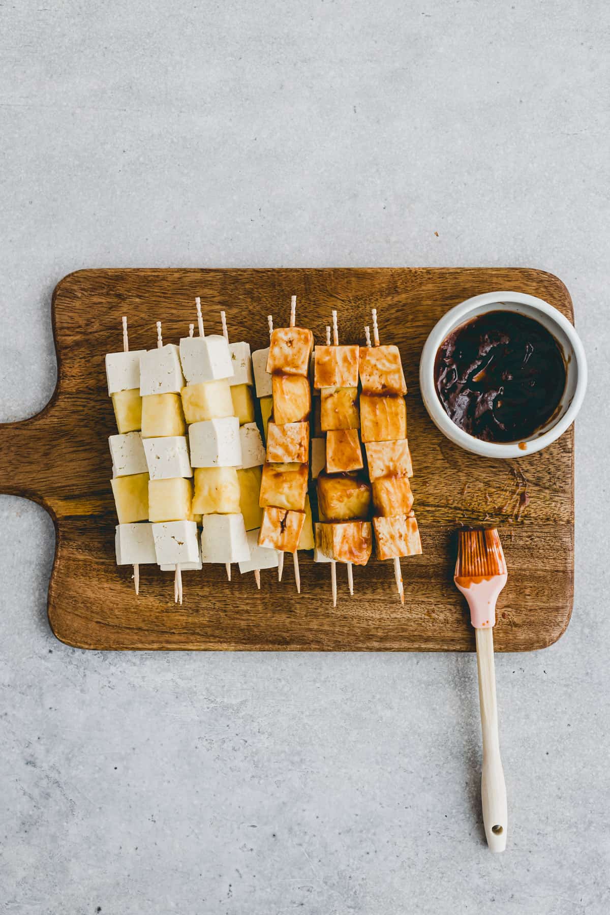 Top view of a chopping board with assembled tofu skewers being costed with BBQ sauce. 