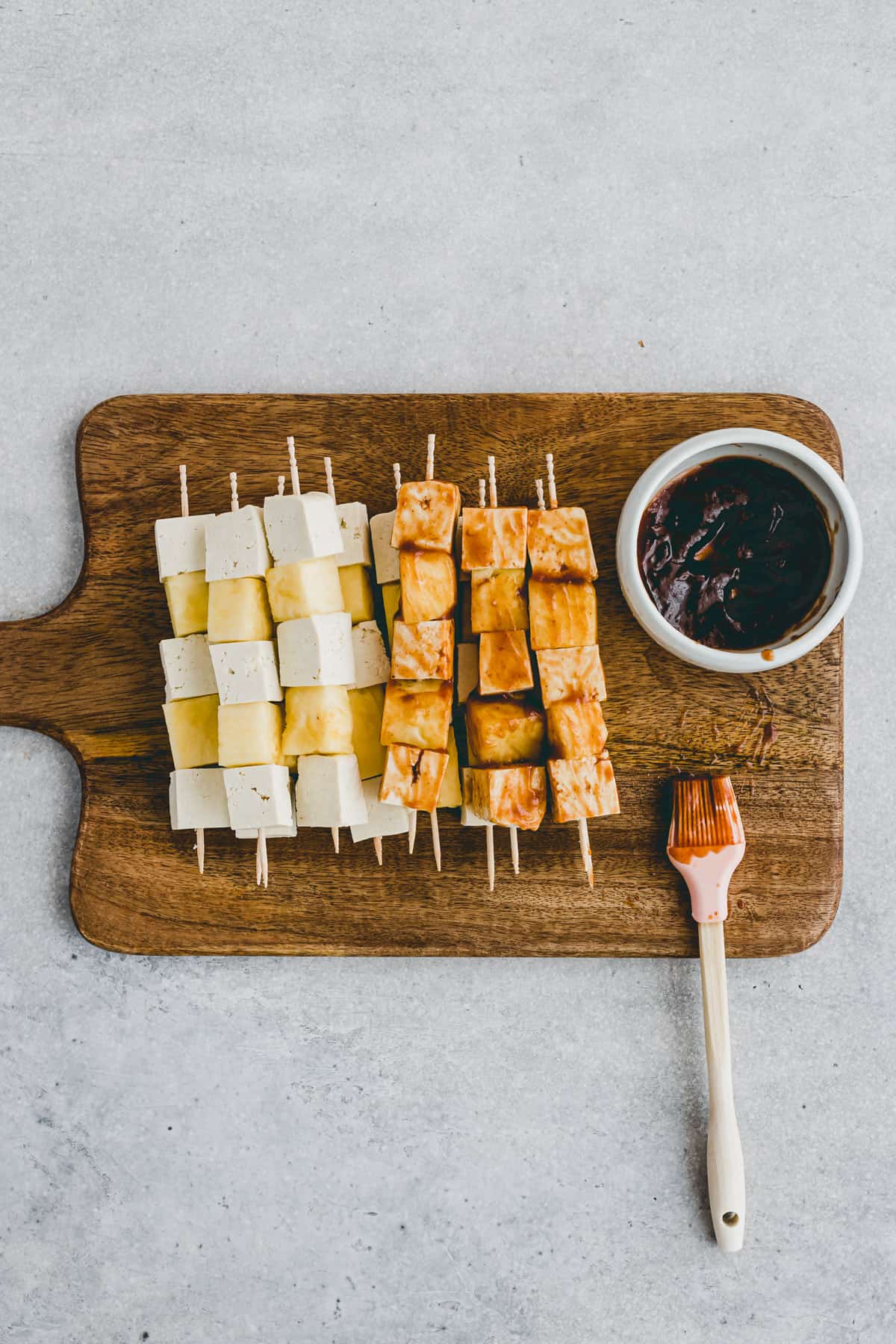 Top view of a chopping board with assembled tofu skewers being costed with BBQ sauce.