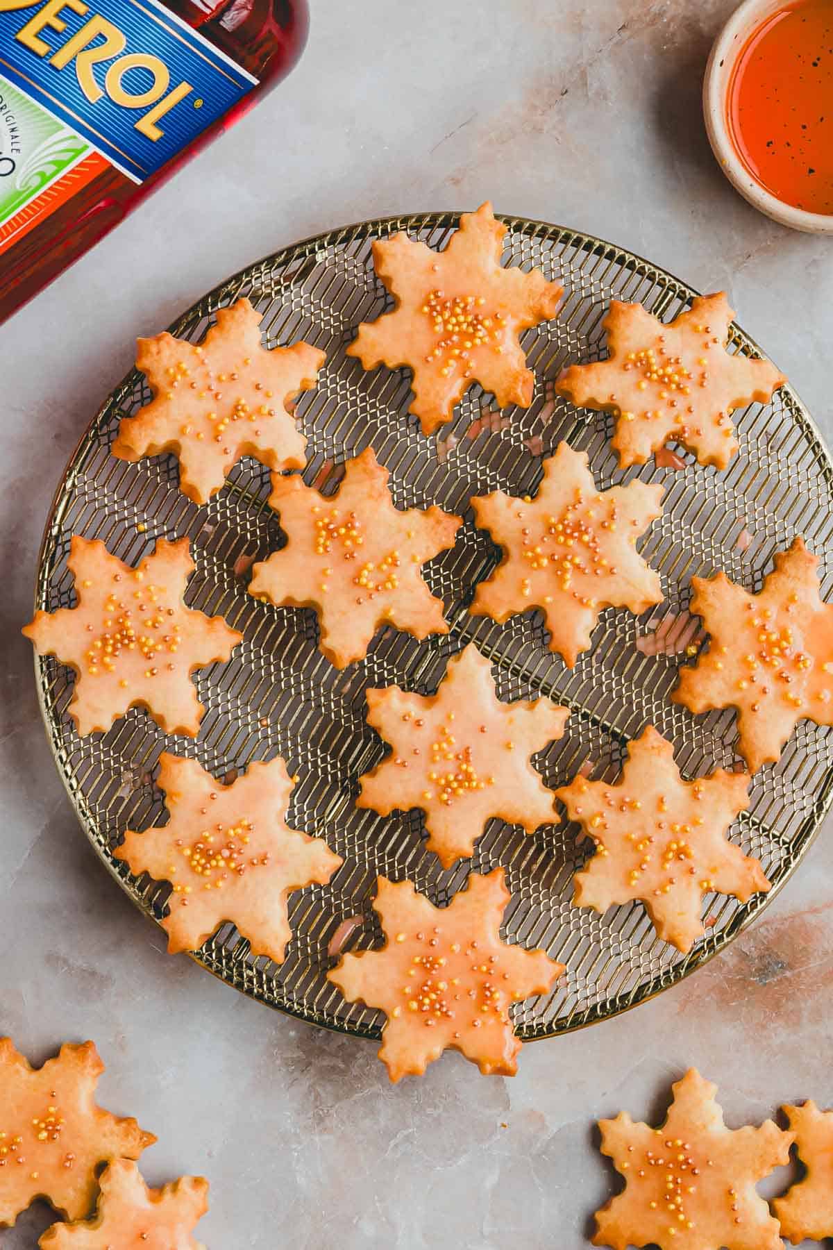 Aperol Plätzchen mit streudekor auf einem goldenen auskühlgitter neben einer flasche aperol